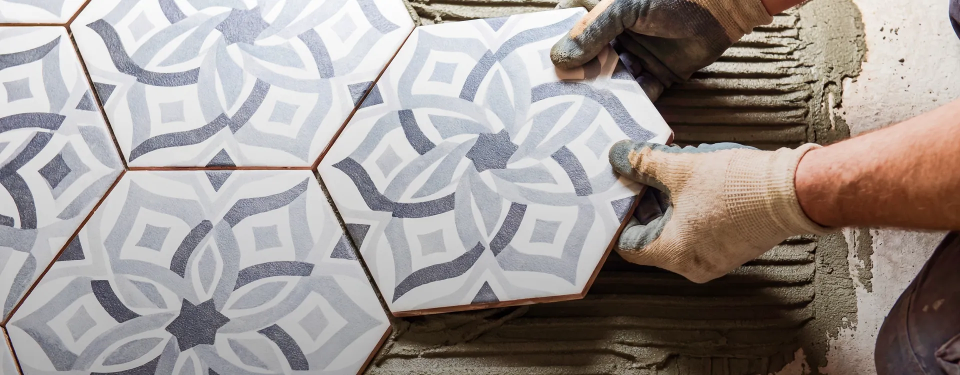Close-up of a worker wearing protective gloves placing a white, gray, and dark blue patterned hexagonal tile onto a layer of combed thin-set mortar during floor installation.